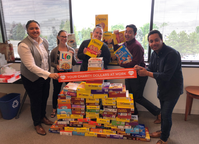 A photo of several people and a lot of food collected for a food drive.  The people are holding a sign that reads: your charity dollars at work.